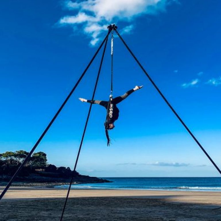 An acrobat hangs from a triangular frame whilst doing the splits upside down on a beautifully sunny day.