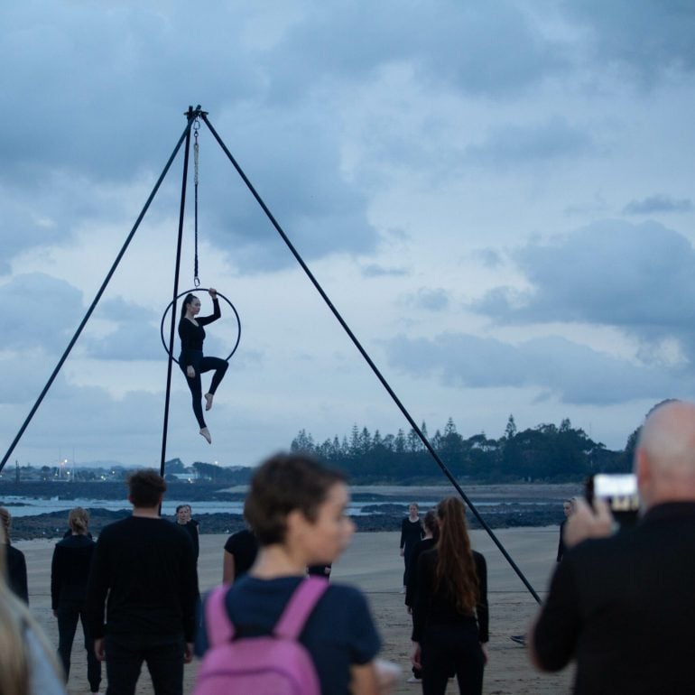 Image of mapali, 2019, a woman hangs in a hoolahoop high in the air above the onlooking crowd at dawn.