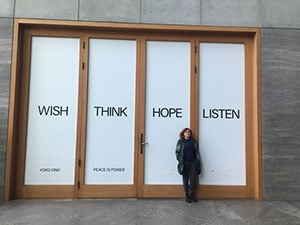 Lindy in front of large doors with writing on them from Yoko Ono’s Peace is Power, Wish. Think. Hope. Listen.