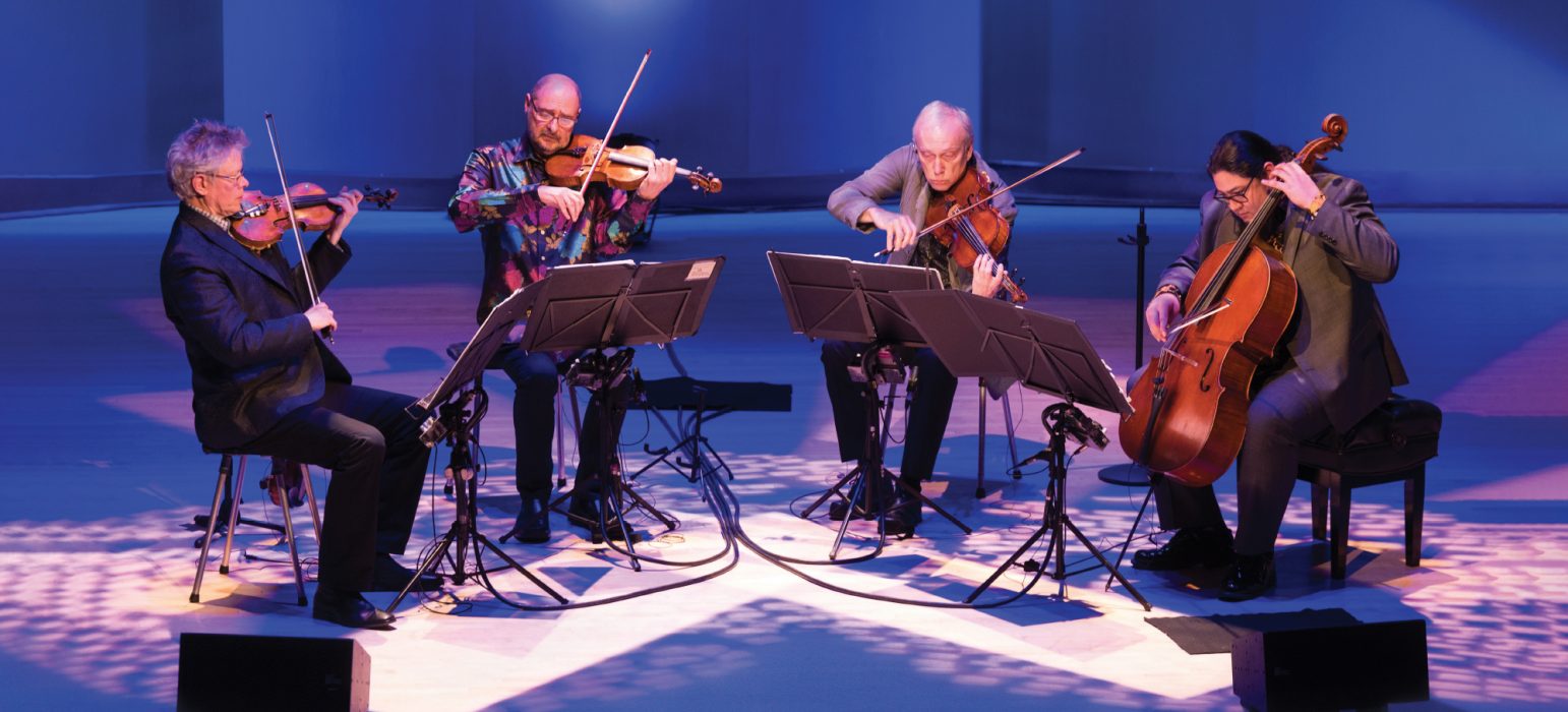 Kronos Quartet hero image, four performers sit under clue/purple light on stage, two violinists, one violist and a cellist all performing