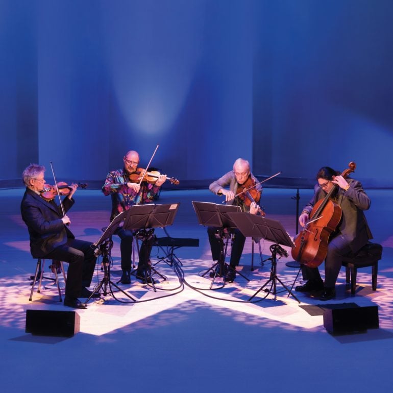 KRONOS QUARTET, four people sit in a semicircle on stage under blue/purple light, engrossed in their performance we see them looking directly towards their sheet music and performing stringed instruments.