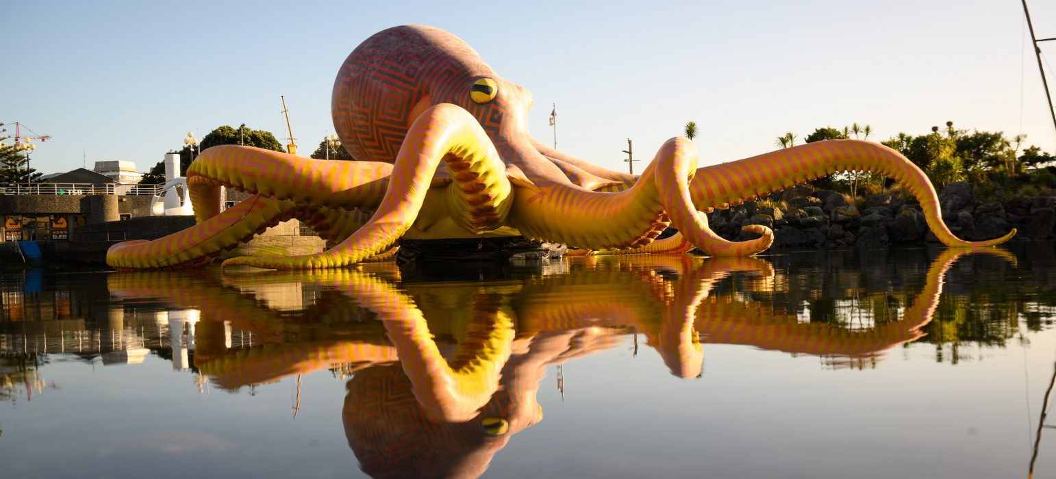Image description: An enormous inflatable octopus sits above a body of water. The octopus is yellow and orange with Maori markings across its curly tentacles and bulging mantle.