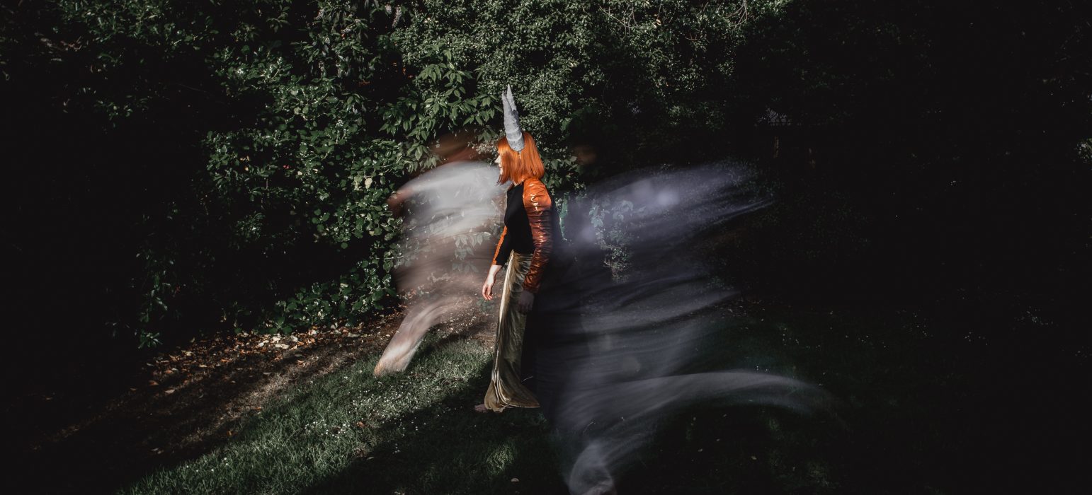 A person with shoulder-length red hair and pointed grey horns is walking through a patch of green grass and shrubbery. Surrounding the person is a blur of action and movement.
