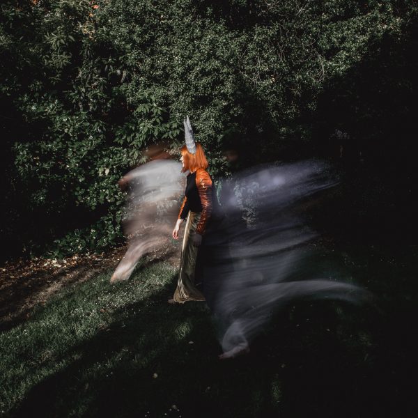 A person with shoulder-length red hair and pointed grey horns is walking through a patch of green grass and shrubbery. Surrounding the person is a blur of action and movement.