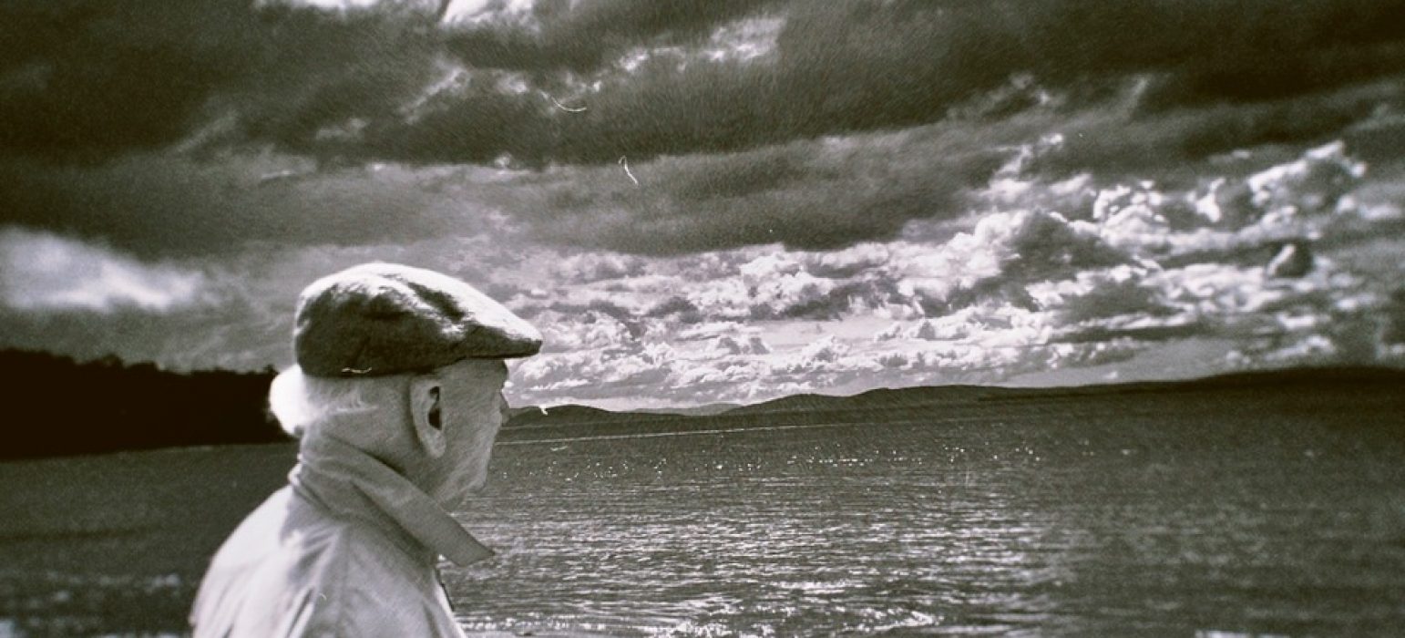 A black and white 120mm negative of an elderly person looking out to the horizon of a body of water. In the distance are hills and above is a cloudy sky with sun rays emerging throughout.