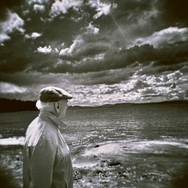 A black and white 120mm negative of an elderly person looking out to the horizon of a body of water. In the distance are hills and above is a cloudy sky with sun rays emerging throughout.