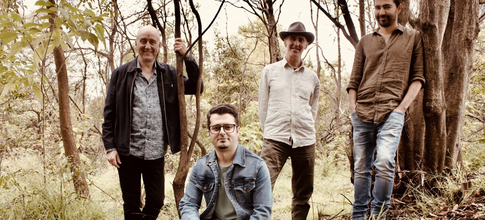 Four people (Hamed Sadeghi and Band) stand and kneel amongst thin trees that have lost their leaves. All people are wearing a mix of jeans, button-down shirts, boots and felt hats in earthy colours. All people are smiling at the camera.