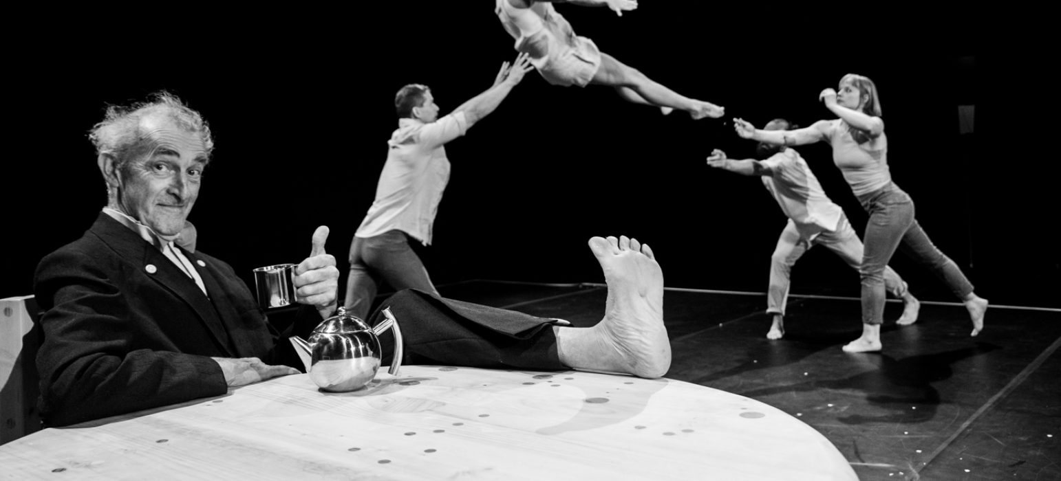 An acrobat is thrown into the air in the background. In the foreground a person (Tony Rooke) drinks a cup of tea with their feet propped up on a table.