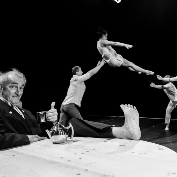 An acrobat is thrown into the air in the background. In the foreground a person (Tony Rooke) drinks a cup of tea with their feet propped up on a table.