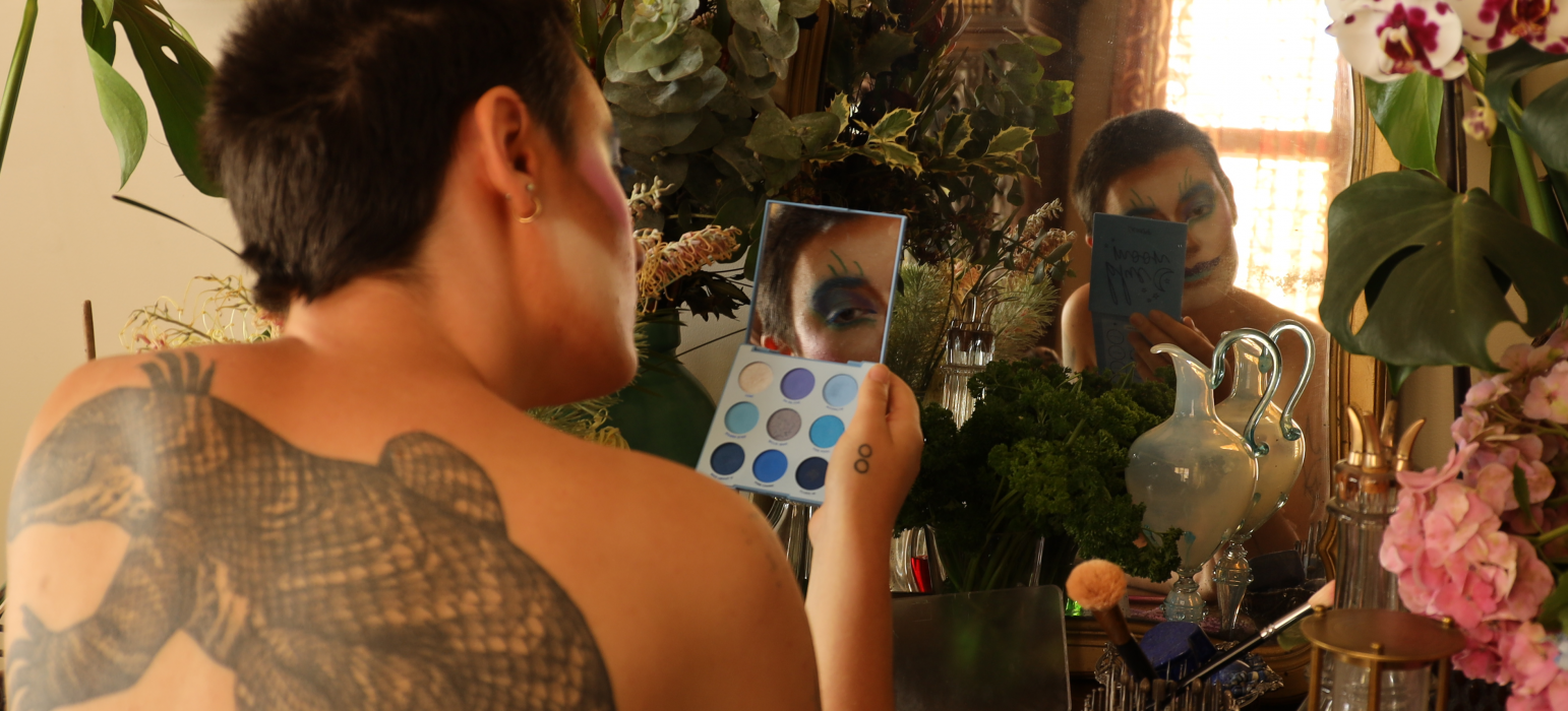 A person sits surrounded by plants and ornaments while facing a mirror. The person inspect their colourful makeup while the camera show's their bare back which is almost covered by a large tattoo of a lizard.