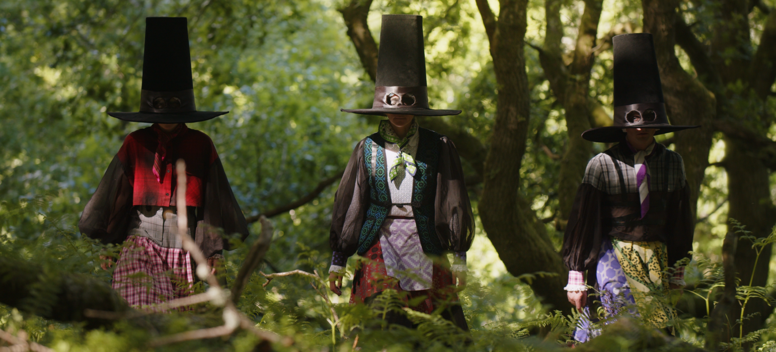 Image description: Three people in tall black hats stand amongst mossy tress and ferns. The three people's faces are covered by their hats, with holes provided for their eyes. They wear layered outfits with puffed sleeves and geometric patterns.