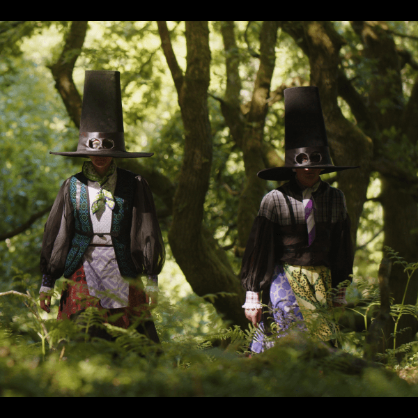 Image description: Three people in tall black hats stand amongst mossy tress and ferns. The three people's faces are covered by their hats, with holes provided for their eyes. They wear layered outfits with puffed sleeves and geometric patterns.