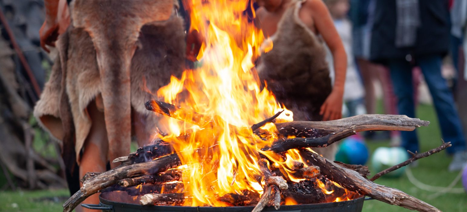 Image description: A large bundle of wood in ablaze in the centre of the picture. Surrounding the fire are various people wearing animal skins and body paint.