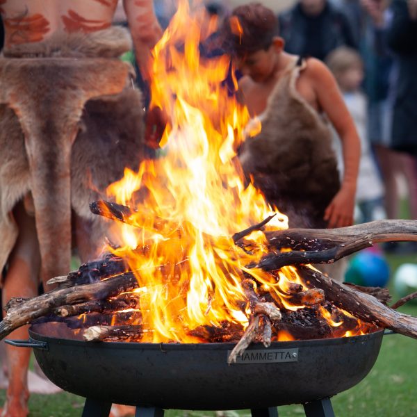 Image description: A large bundle of wood in ablaze in the centre of the picture. Surrounding the fire are various people wearing animal skins and body paint.