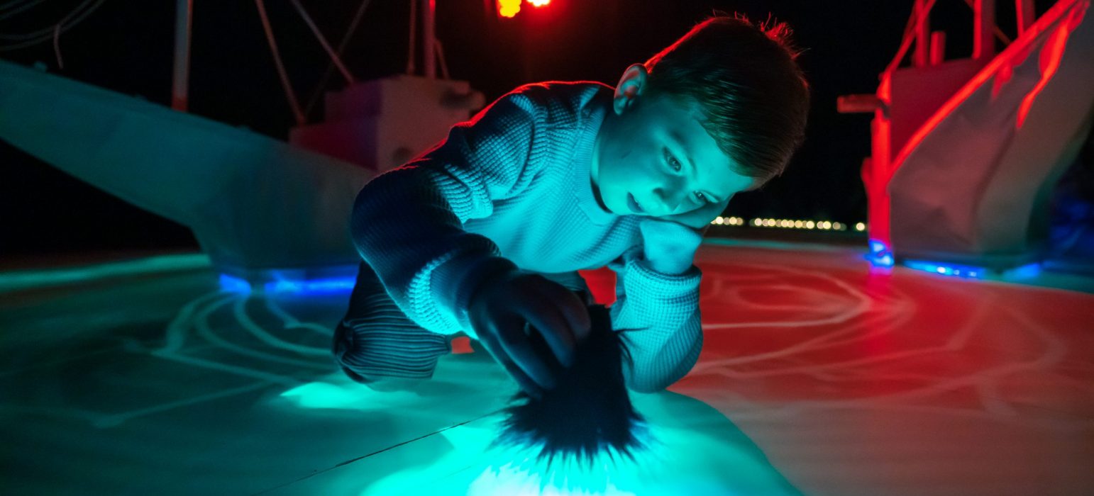 A child is kneeling on the floor while holding a fluffy paintbrush. The paintbrush is drawing bright light patterns on the floor, illuminating the surroundings.