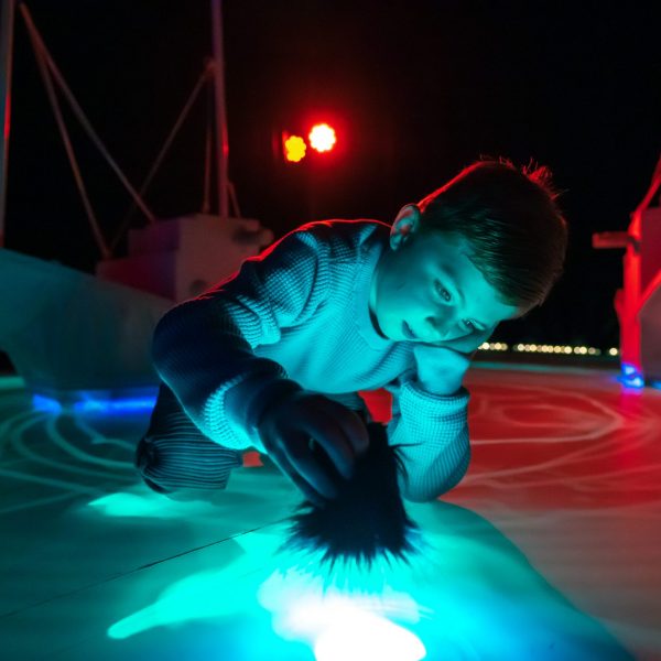 A child is kneeling on the floor while holding a fluffy paintbrush. The paintbrush is drawing bright light patterns on the floor, illuminating the surroundings.