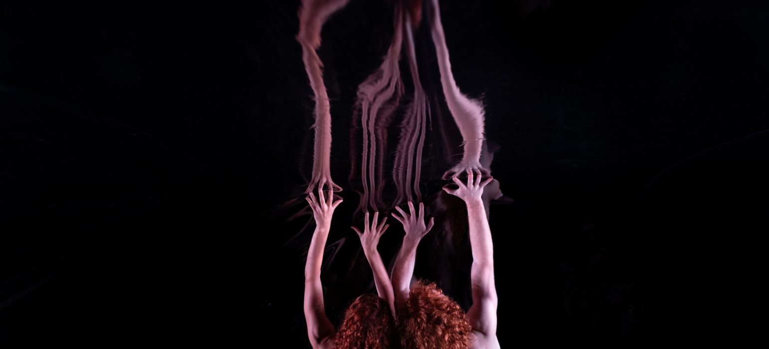 Two people with bright, curly red hair and bare backs stretch their arms above their heads, reaching into their dark and unknown surroundings.