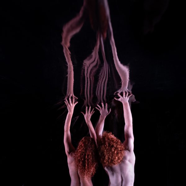 Two people with bright, curly red hair and bare backs stretch their arms above their heads, reaching into their dark and unknown surroundings.