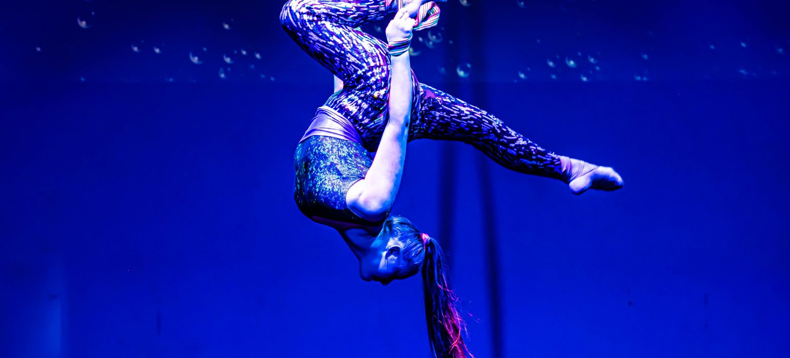 A person wearing patterned spandex hangs upside down while held by silk lengths of fabric.