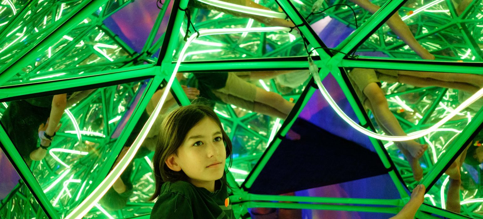 A young person with long dark hair sits on the floor and looks up to the inside of a mirrored dome.