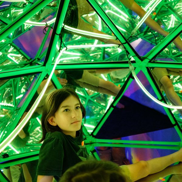 A young person with long dark hair sits on the floor and looks up to the inside of a mirrored dome.