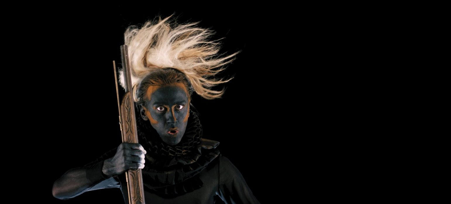 A person stares at the camera with wide eyes, a painted face and parted lips as if to make a deep sound. The person holds the barrel of a rifle. They wear a period ruffled collar and a traditional headdress.