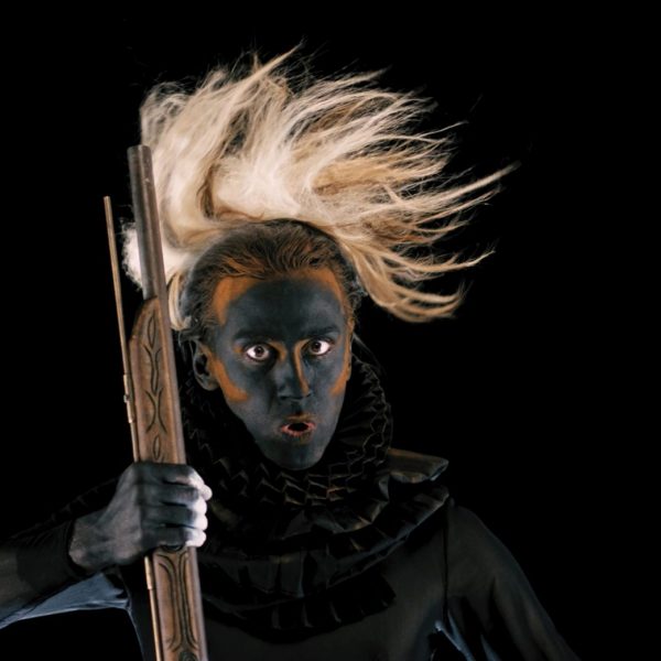 A person stares at the camera with wide eyes, a painted face and parted lips as if to make a deep sound. The person holds the barrel of a rifle. They wear a period ruffled collar and a traditional headdress.