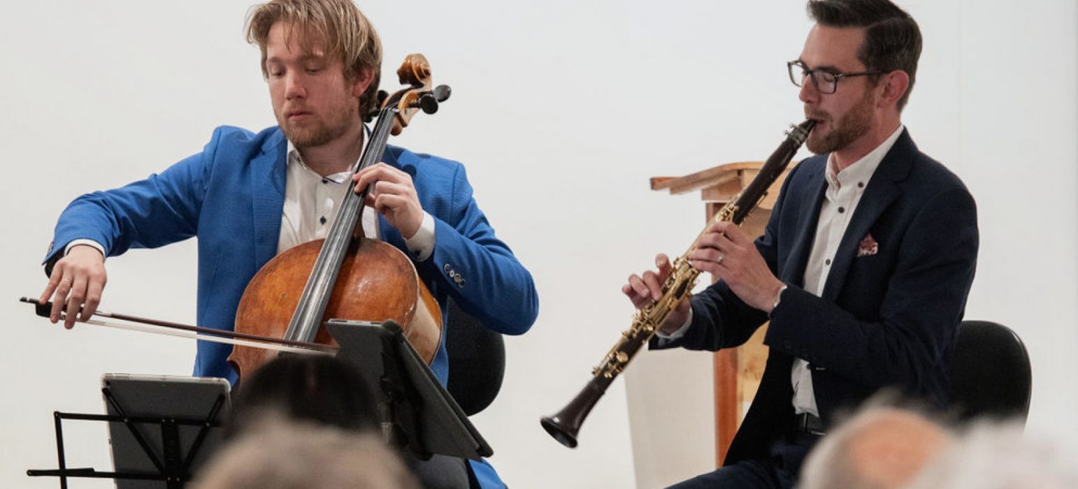 A person playing the cello sits beside a person playing the clarinet. Both people wear suits and are focused on their sheet music.