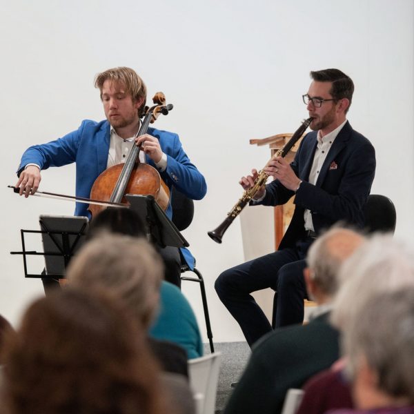 A person playing the cello sits beside a person playing the clarinet. Both people wear suits and are focused on their sheet music.