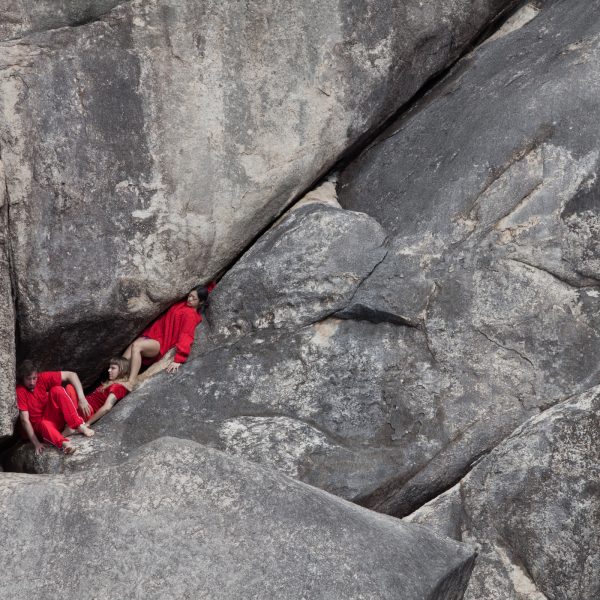 Three people in red clothing are wedged in the cracks of a rock face. The people use each others bodies to balance their position in the rocks.