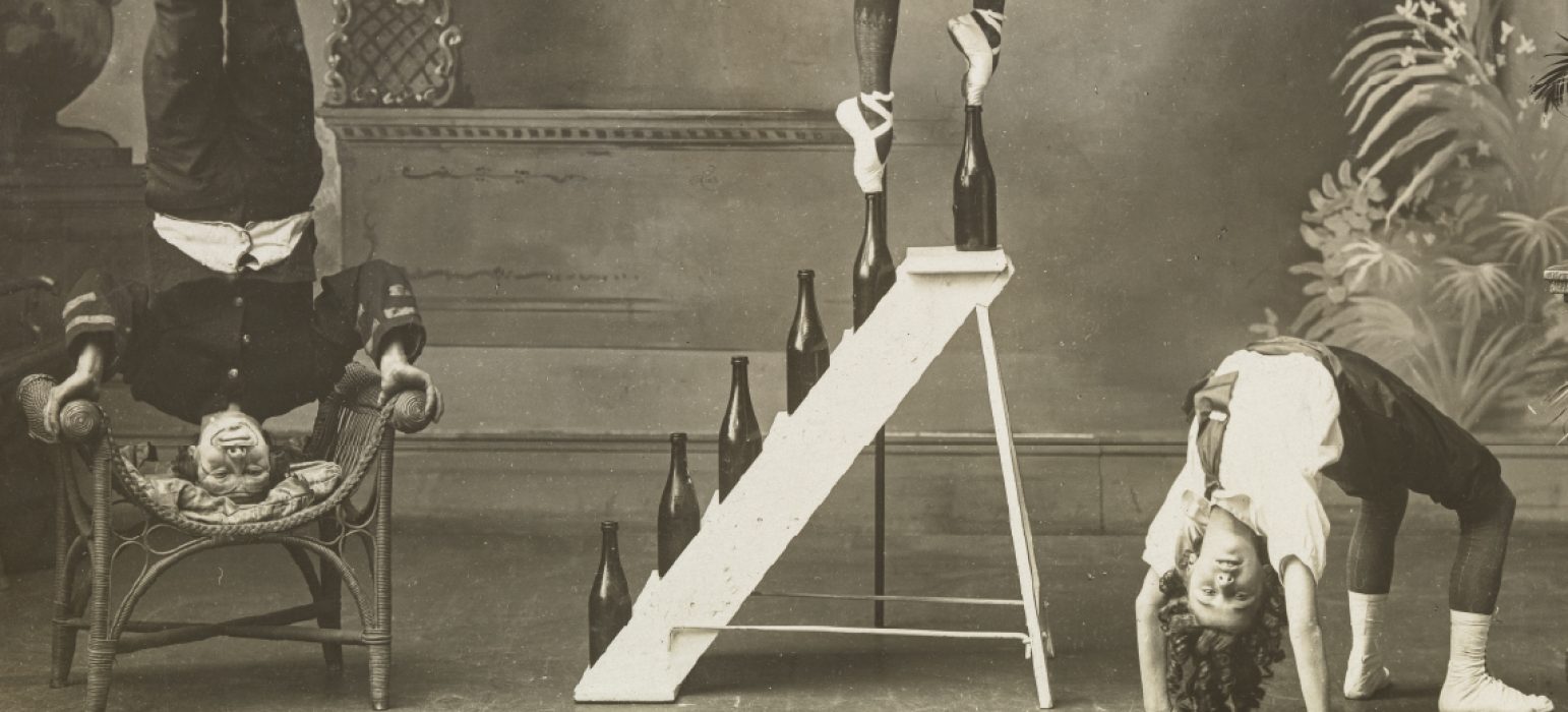A black and white picture featuring a performer wearing ballet pointe shoes is tiptoeing on bottle tops set on consecutive steps of a ladder, another performer is standing on their head and another is arching their back to plant both feet and palms on the floor.