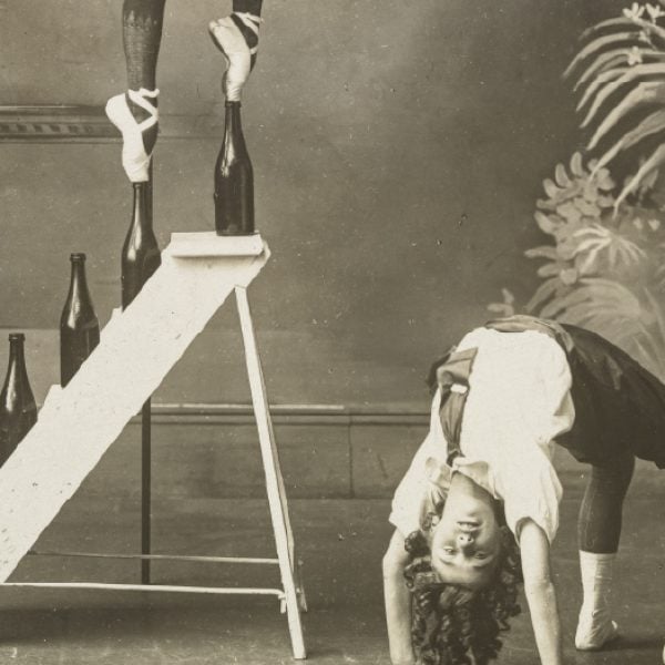 A black and white picture featuring a performer wearing ballet pointe shoes is tiptoeing on bottle tops set on consecutive steps of a ladder, another performer is standing on their head and another is arching their back to plant both feet and palms on the floor.