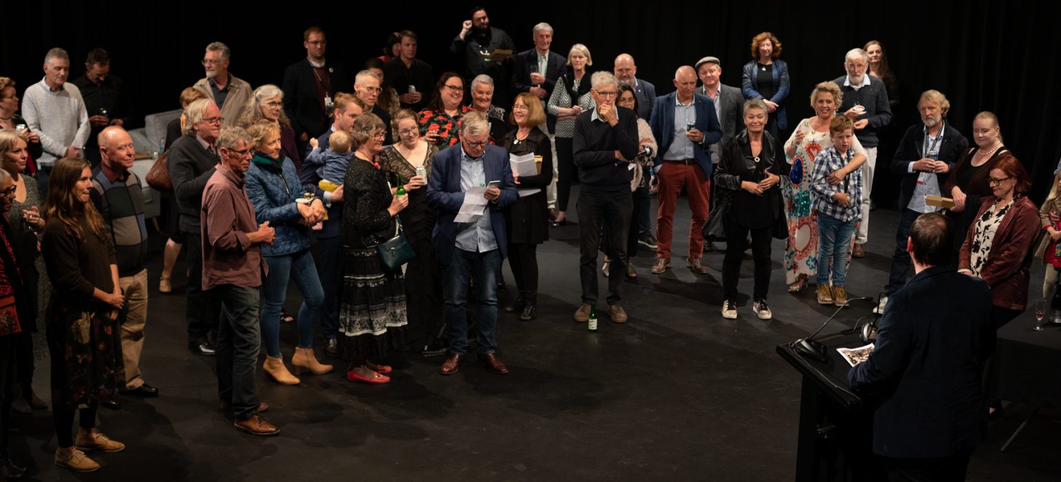 A semicircle of crowded people are listening to a speaker standing at a plinth. Amongst the crowd are various member of the Ten Days team and friends who are smiling.