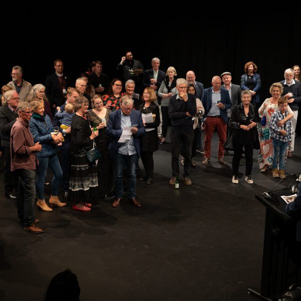 A semicircle of crowded people are listening to a speaker standing at a plinth. Amongst the crowd are various member of the Ten Days team and friends who are smiling.
