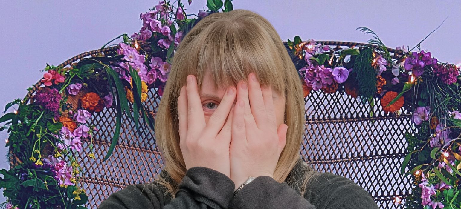 A person sits atop a floral throne. They cover their face, peeking one eye out between their fingers.