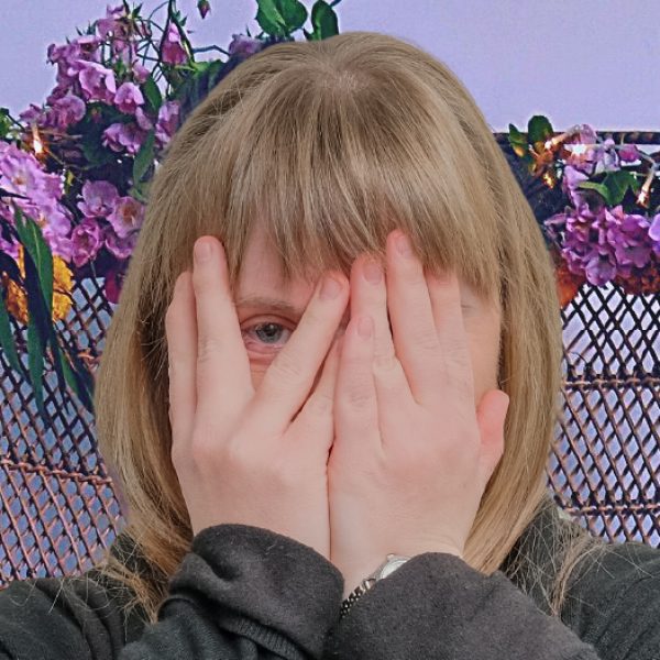 A person sits atop a floral throne. They cover their face, peeking one eye out between their fingers.