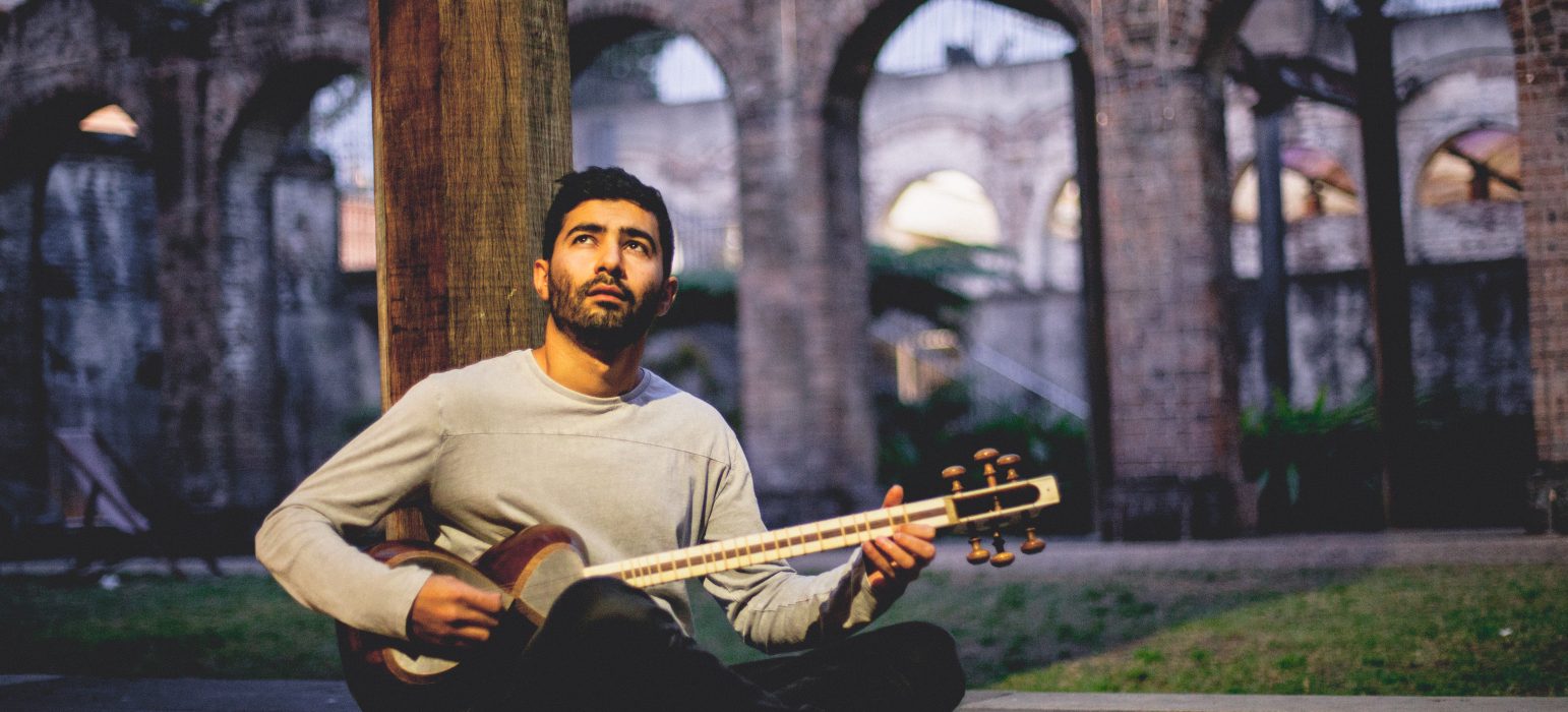 A person (Hamed Sadeghi) sits on the ground with crossed legs. Hamed looks to the sky as he plays a stringed, guitar-like instrument.