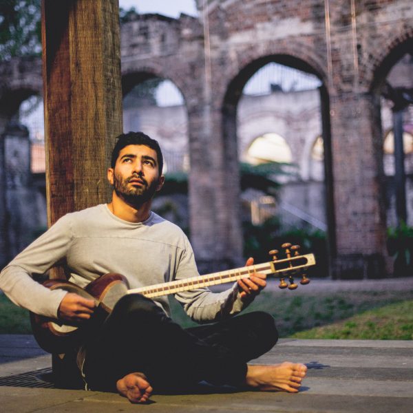 A person (Hamed Sadeghi) sits on the ground with crossed legs. Hamed looks to the sky as he plays a stringed, guitar-like instrument.