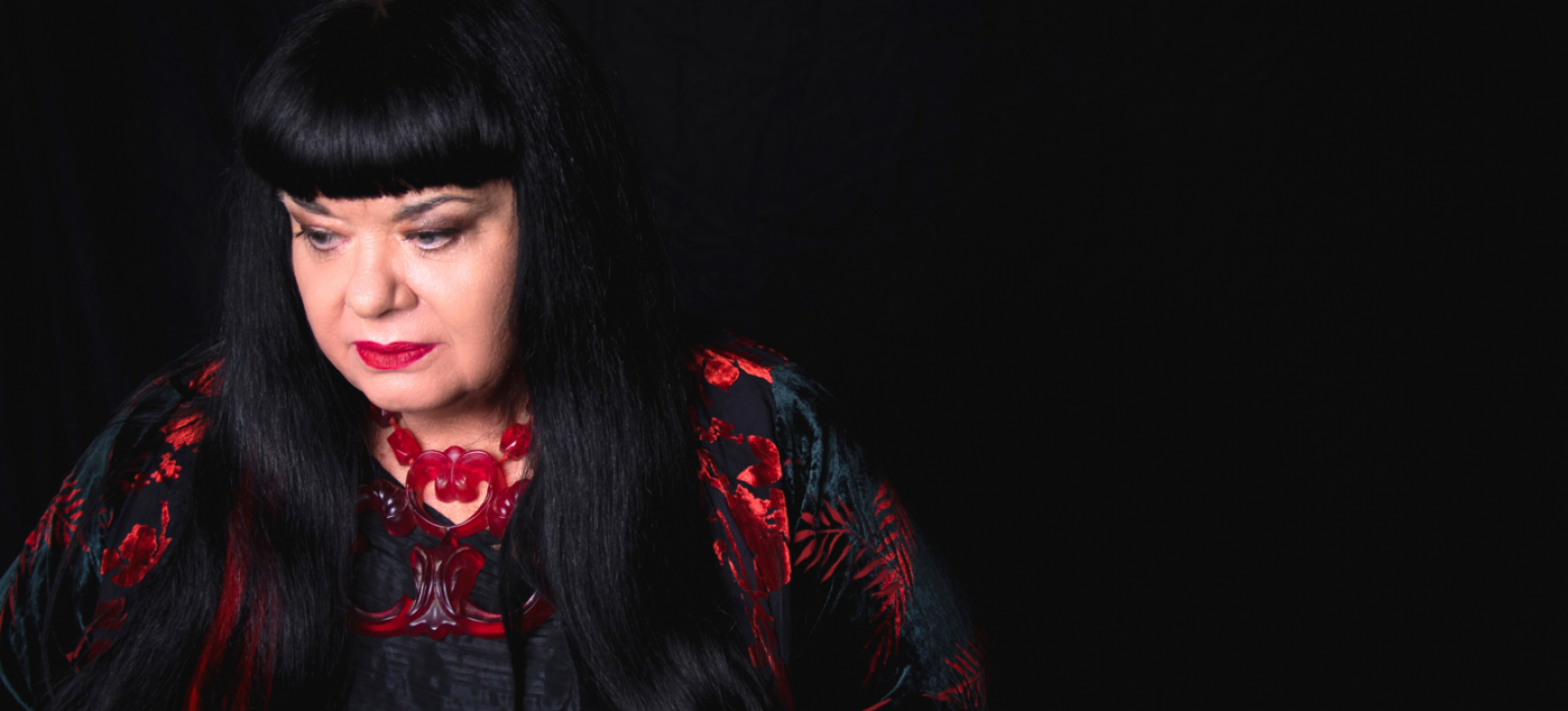 A person (Lynette Wallworth) with long black hair and a blunt fringe looks off into the distance. Contrasting with a black background and clothing, this person wears bright red lipstick, a red necklace and a red pattern on their dress.