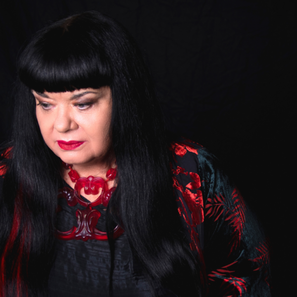 A person (Lynette Wallworth) with long black hair and a blunt fringe looks off into the distance. Contrasting with a black background and clothing, this person wears bright red lipstick, a red necklace and a red pattern on their dress.