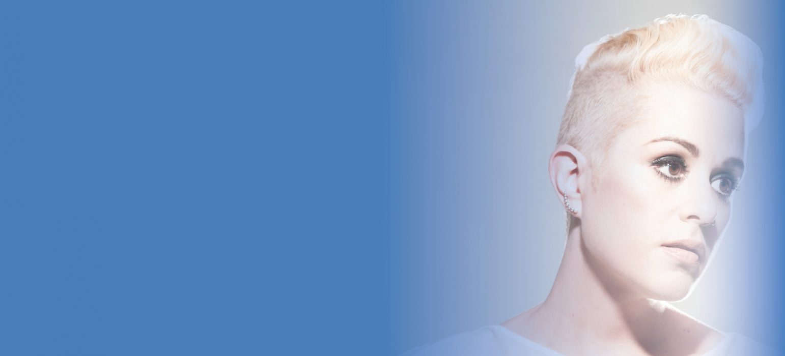 A person (Katie Noonan) with short blonde hair and wearing a white gown. Illuminated by a beacon of light and atop of rich blue background, Katie glows as she looks off into the distance.