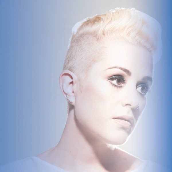 A person (Katie Noonan) with short blonde hair and wearing a white gown. Illuminated by a beacon of light and atop of rich blue background, Katie glows as she looks off into the distance.