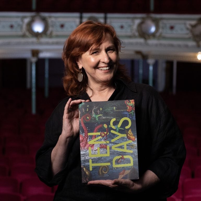 Image of Dr Lindy Hume AM standing on a stage holding the Ten Days on the Island 2023 brochure