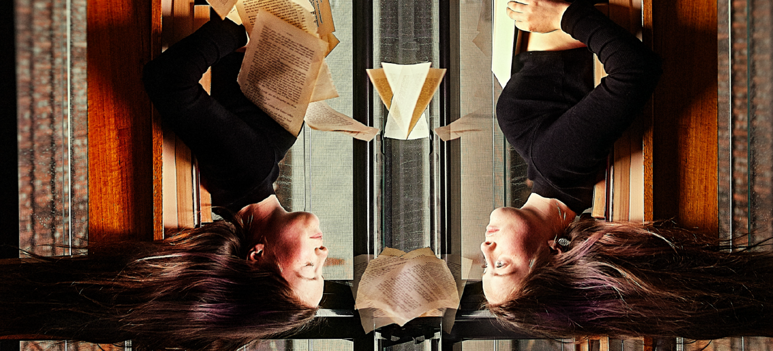 A person is upside down, facing a version of themselves like a mirror. Wind blows the person's hair away from their face and sends book pages flying around the image.