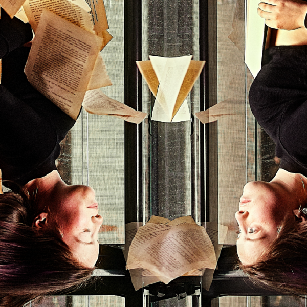 A person is upside down, facing a version of themselves like a mirror. Wind blows the person's hair away from their face and sends book pages flying around the image.