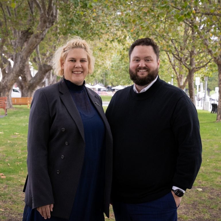 Two people stand in a park land area, with trees either side.