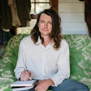 Ha! Fair. Let me look properly. Portrait of writer Bert Spinks, seated on a bold green floral couch in a rustic timber cabin, pen poised over an open notebook in his lap, long curly hair framing a direct and thoughtful gaze.
