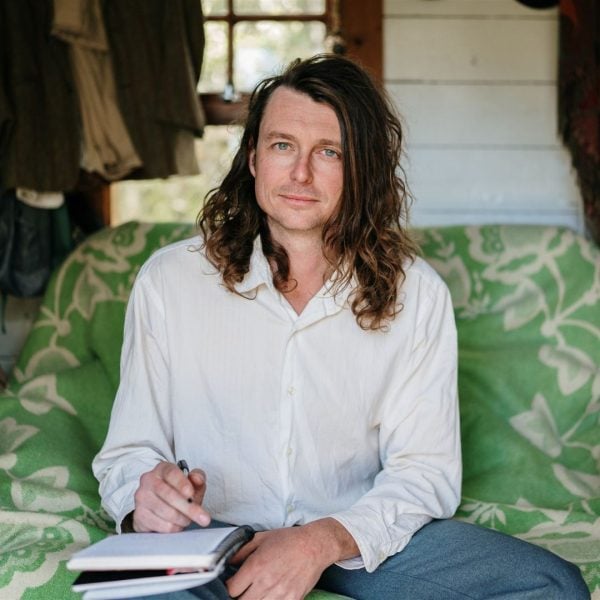 Ha! Fair. Let me look properly. Portrait of writer Bert Spinks, seated on a bold green floral couch in a rustic timber cabin, pen poised over an open notebook in his lap, long curly hair framing a direct and thoughtful gaze.