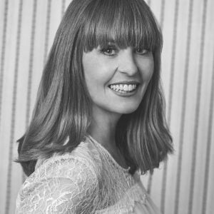 Black and white portrait of Kirsten Galliott, smiling over her shoulder with a blunt fringe, shoulder-length hair, and a delicate lace top against a softly striped background.